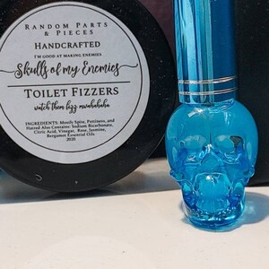 May include: A blue glass skull-shaped bottle with a silver cap. The bottle is filled with a clear liquid. The label on the bottle reads "Skulls of my Enemies Toilet Fizzers watch them fizz mwahahaha".