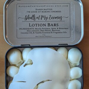 May include: A metal tin containing lotion bars shaped like skulls. The tin lid has text that reads "Skulls of My Enemies" and lists ingredients. The lotion bars are white and the tin is open, revealing the contents.