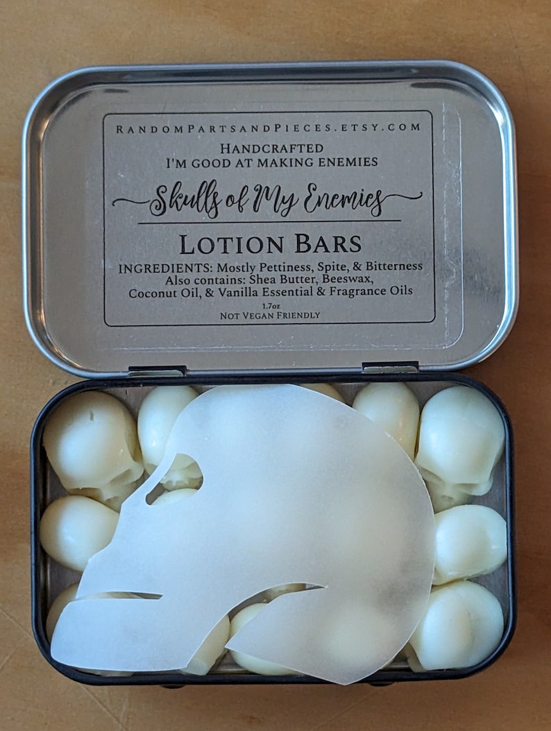 May include: A metal tin containing lotion bars shaped like skulls. The tin lid has text that reads "Skulls of My Enemies" and lists ingredients. The lotion bars are white and the tin is open, revealing the contents.