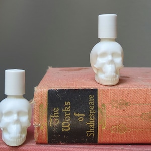 May include: Two white skull-shaped glass bottles with white caps. The bottles are sitting on top of a red book with the title "The Works of Shakespeare" printed on the spine.