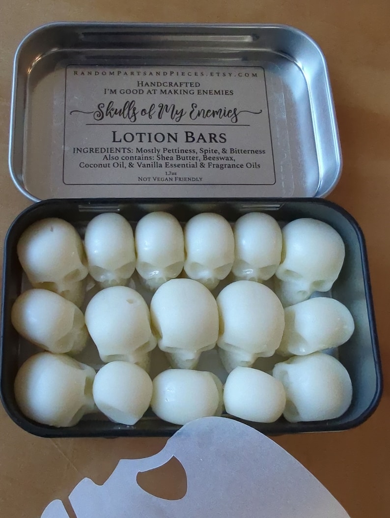 May include: An open metal tin filled with white skull-shaped lotion bars. The tin's label reads "Skulls of My Enemies" and lists ingredients like shea butter and coconut oil. The bars are arranged neatly inside the tin.