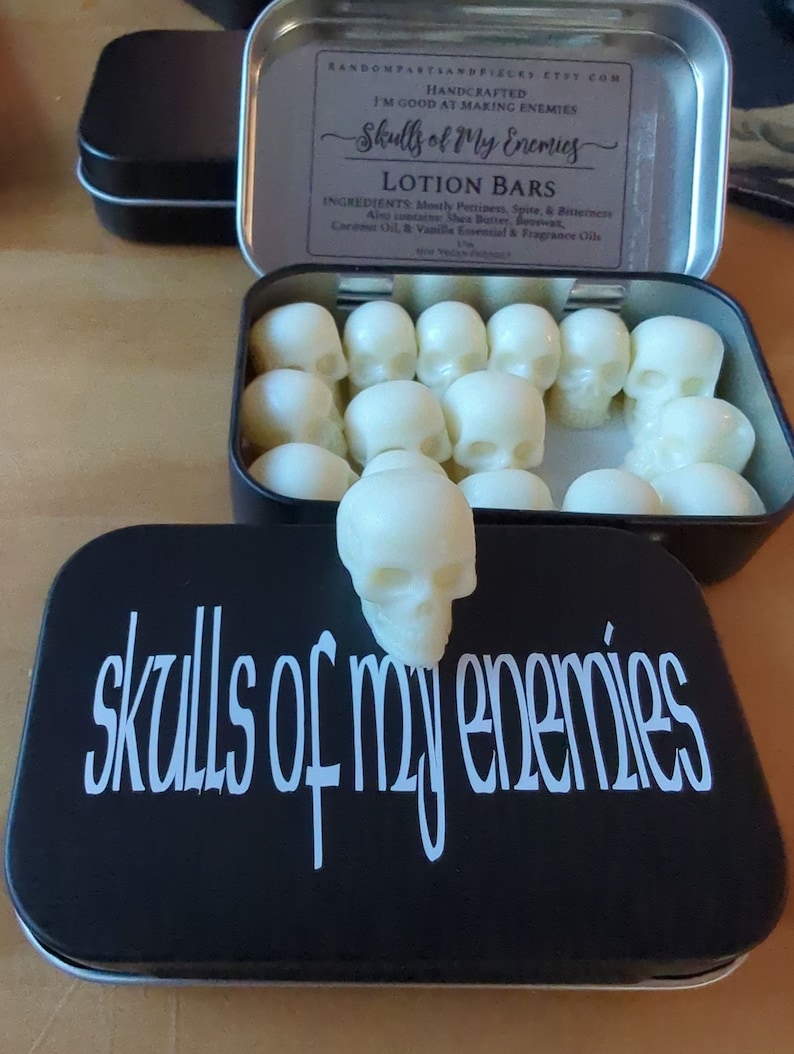 May include: A black tin with the words "skulls of my enemies" in white script. Inside, skull-shaped lotion bars are visible. The tin is open, revealing more bars. The top of the tin reads "Skulls of My Enemies" and lists ingredients.