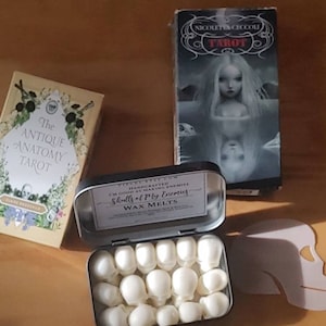 May include: An open tin of white skull-shaped wax melts, labeled "Skulls of My Enemies." Beside it are two tarot card boxes, one titled "The Antique Anatomy Tarot" and the other "Nicoletta Ceccoli Tarot."