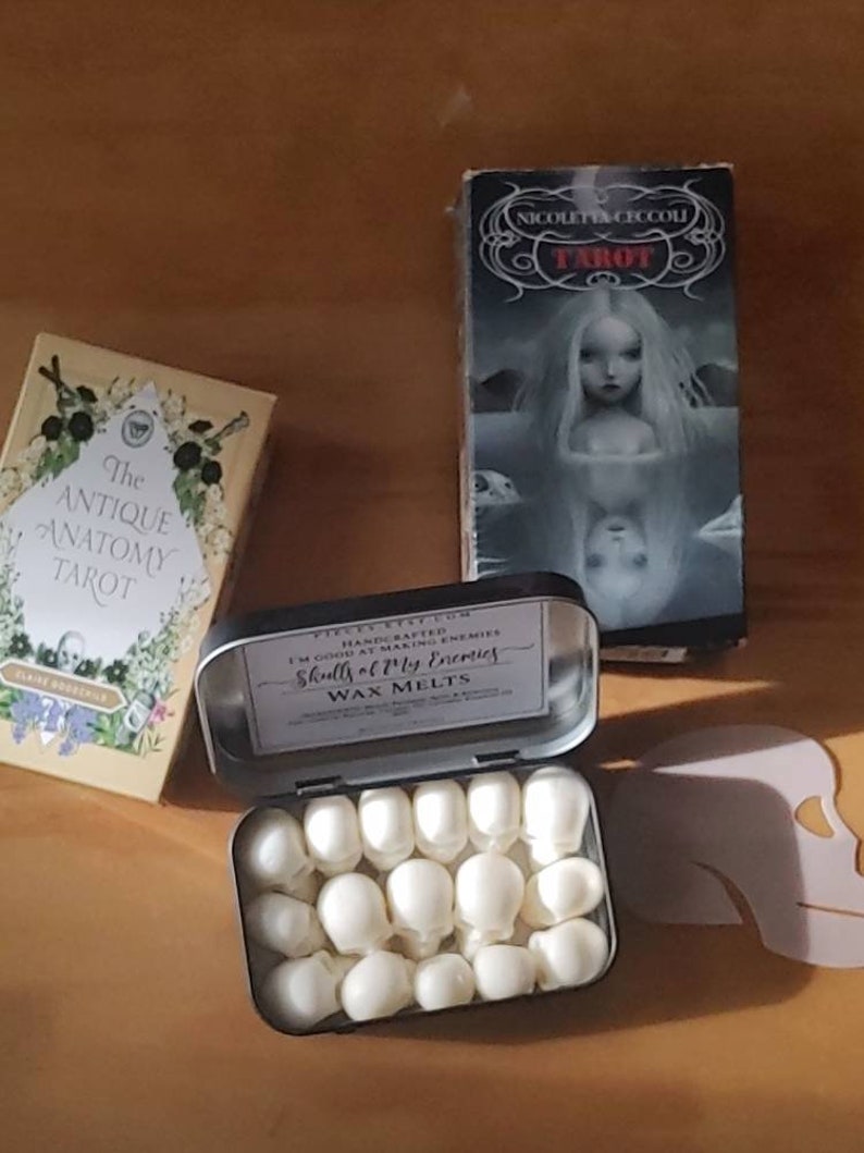 May include: An open tin of white skull-shaped wax melts, labeled "Skulls of My Enemies." Beside it are two tarot card boxes, one titled "The Antique Anatomy Tarot" and the other "Nicoletta Ceccoli Tarot."