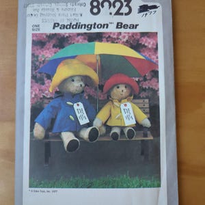 May include: Simplicity 8923 sewing pattern featuring Paddington Bear. The pattern cover shows two Paddington Bear plush toys wearing blue and yellow raincoats, hats, and holding an umbrella. The background is a pink floral design.