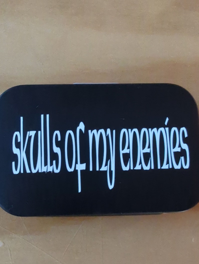 May include: A black rectangular object with the words "skulls of my enemies" in white, stylized cursive font. The text is centered on the object.