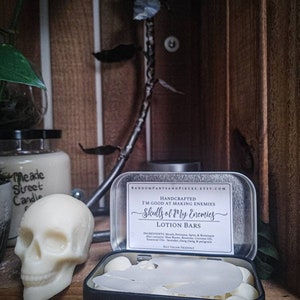 May include: A white skull-shaped candle and a black tin containing lotion bars. The tin has a label that reads "Skulls of My Enemies" and "Lotion Bars". A metal rose and a jar of candles are in the background.