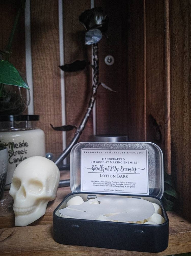 May include: A white skull-shaped candle and a black tin containing lotion bars. The tin has a label that reads "Skulls of My Enemies" and "Lotion Bars". A metal rose and a jar of candles are in the background.