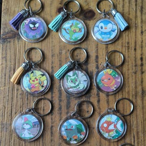 May include: A collection of twelve circular acrylic keychains, each featuring a different colorful cartoon creature illustration. Each keychain includes a metal split ring and a small decorative tassel in various colors like purple, teal, and yellow.