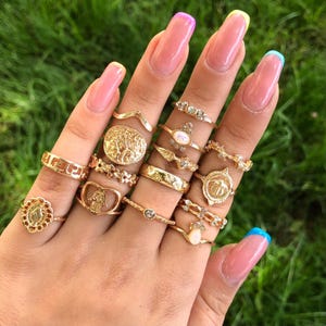 May include: A collection of gold-toned metal rings worn on fingers, featuring various designs including a hamsa hand, a circular coin-style pendant, a Greek key pattern, floral motifs, small clear crystals, and oval-shaped cabochon stones.