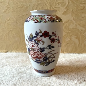 May include: A white ceramic vase with a floral design. The vase has a gold rim and is decorated with red, blue, and green flowers.