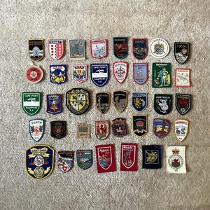 Può includere: Una collezione di toppe ricamate, con vari disegni e testo. Le toppe sono a forma di scudo e circolare, con colori vivaci e immagini dettagliate. Alcune toppe mostrano nomi di città, stemmi e loghi. Le toppe sono disposte su una superficie di colore neutro.