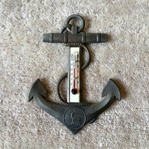 May include: A metallic anchor-shaped wall thermometer with a dark grey finish. The center features a vertical glass tube with red liquid and temperature markings in Celsius and Fahrenheit. A circular emblem at the base depicts a standing deer.