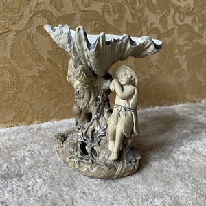 Peut inclure: Figurine décorative représentant un bol en forme de coquillage soutenu par une base sculptée avec un enfant endormi. La base a un aspect texturé et vieilli avec une palette de couleurs neutres beige, grise et blanche.