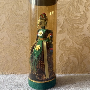 Pode incluir: Uma boneca decorativa dentro de um tubo cilíndrico de plástico transparente com base verde. A boneca apresenta um rosto pintado, um corpete verde com detalhes de flores douradas e uma saia castanha estampada. Usa um toucado e contas coloridas.