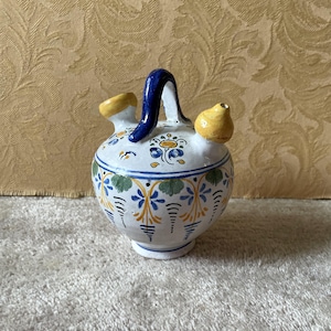 Può includere: Brocca in ceramica bianca con manico e beccuccio blu, e beccuccio ed elemento decorativo gialli. La brocca è adornata con motivi floreali blu, gialli e verdi. La brocca è alta circa 15 cm.