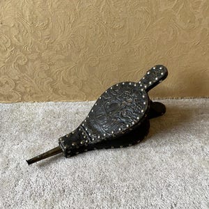 Puede incluir: Fuelle antiguo de cuero negro con detalles en relieve y tachuelas plateadas. El fuelle presenta una boquilla metálica larga y un patrón decorativo. El diseño sugiere un estilo vintage o histórico.