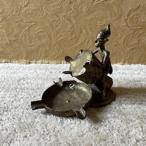 Puede incluir: Un juego de ceniceros de bronce con una figura sentada que sostiene una tapa en forma de tortuga. El cenicero tiene dos cuencos separados con una tapa en forma de tortuga. El juego es de bronce y tiene un aspecto vintage.