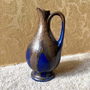 Pode incluir: Um jarro de cerâmica com corpo bulboso, pescoço estreito e uma única alça. A superfície apresenta um esmalte azul cobalto brilhante contrastado com um padrão de gotejamento texturizado em bronze acastanhado metálico que desce da borda.