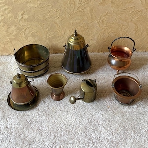 Op de afbeelding: Een verzameling miniatuur messing en koperen items, waaronder een emmer, een klokvormige container, een kleine ketel, een gieter en een kleine beker. De items zijn gerangschikt op een wit oppervlak.