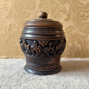 Può includere: Un vaso di legno marrone scuro con coperchio e un motivo floreale intagliato al centro. Il vaso ha una base rotonda e un piccolo pomello sul coperchio. La venatura del legno è visibile, conferendogli un aspetto rustico. Alto circa 15 cm.