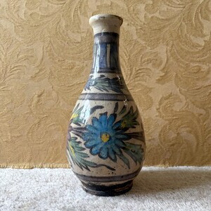 Può includere: Un vaso in ceramica dipinto a mano con collo stretto e corpo bulboso. Il vaso presenta un motivo floreale blu con foglie verdi e accenti gialli su sfondo color crema. Il vaso è alto circa 20 cm.