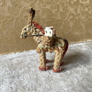 Può includere: Una statuetta di asino fatta a mano con fibre naturali intrecciate, con criniera e coda rosse e marroni e un piccolo vaso bianco sulla schiena. L'asino è adornato con accenti verdi e gialli.