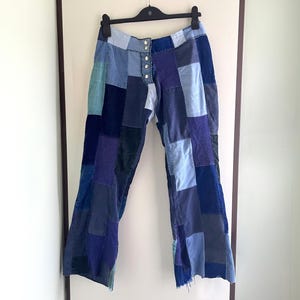 May include: Patchwork pants featuring a variety of blue and indigo fabric squares stitched together. The trousers have a button-up fly with silver-toned metal buttons and are displayed on a black plastic hanger against a plain background.