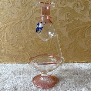 Op de afbeelding: Een heldere glazen parfumfles met roze accenten en een blauw bloemenpatroon. De fles heeft een uitlopende bovenkant en een kleine schaalvormige basis, ondersteund door een gebogen steel. De basis is cirkelvormig.