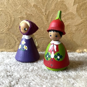 Peut inclure: Deux poupées en bois. L'une porte une robe violette avec des accents de marguerites blanches et un bonnet violet. L'autre porte une robe rouge et verte avec des accents floraux blancs et un chapeau rouge.