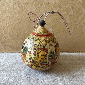 Può includere: Una piccola zucca rotonda dipinta a mano con un coperchio e un manico in corda. La zucca è decorata con motivi floreali e geometrici colorati, e un'immagine centrale di una persona con una corona.