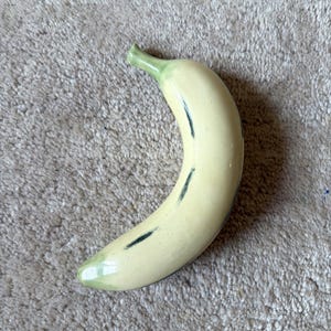 Op de afbeelding: Een keramieken bananenbeeldje met een glanzende lichtgele afwerking, voorzien van een lichtgroene steel en punt, en subtiele donkergroene geschilderde accenten langs het gebogen lichaam om het uiterlijk van rijp fruit na te bootsen.
