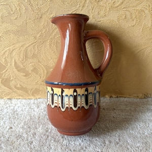 May include: A brown ceramic pitcher with a glossy finish and a visible crackle glaze pattern. The body features a decorative band with a marbled, wavy design in cream, black, and blue tones, accented by a thin dark blue horizontal line above it.
