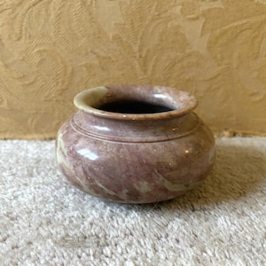 Könnte beinhalten: Eine kleine, runde Vase mit breiter Öffnung. Die Vase besteht aus Stein und zeigt ein wirbelndes Muster aus Rosa und Creme. Der Innenraum ist dunkel, und der Rand ist cremefarben. Geeignet für kleine Blumen.