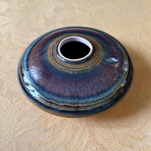 Könnte beinhalten: Eine Keramikvase mit breitem, flachem Boden und einer kleinen, kreisförmigen Öffnung. Die Vase hat eine glänzende Oberfläche mit einer Mischung aus blauen, braunen und violetten Farbtönen. Das Innere der Öffnung ist weiß.