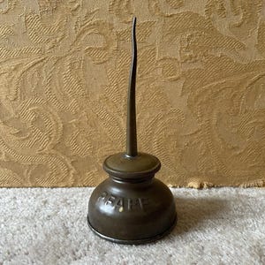 Op de afbeelding: Een vintage metalen oliekan met een lange tuit. De kan is bruin en heeft het woord "PEARE" erop gedrukt.