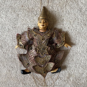 Könnte beinhalten: Handgefertigte Marionettenpuppe in traditioneller Tracht. Die Puppe hat ein detailliertes, verziertes Outfit in Braun-, Gold- und Rottönen, verziert mit Pailletten und Perlen. Die Puppe hat eine konische Kopfbedeckung und bemalte Gesichtszüge. Die Marionette wird an Schnüren aufgehängt.