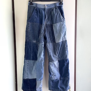 Puede incluir: Pantalones de talle alto confeccionados con varios tonos de tela vaquera azul en un diseño de patchwork. La prenda cuenta con cierre de botón y cremallera, trabillas para cinturón y bolsillos laterales, con un pequeño adorno colgante.