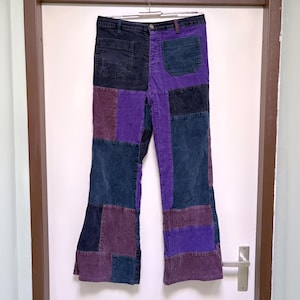 Op de afbeelding: Patchwork broek met een color-block ontwerp in tinten donkerpaars, diep violet en antracietgrijs. Deze broek is voorzien van een knoopsluiting, riemlussen en twee opgestikte voorzakken, opgebouwd uit meerdere stoffen panelen.