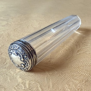 Op de afbeelding: Een heldere glazen parfumfles met een zilverkleurige, sierlijke dop. De fles heeft een cilindrische vorm met verticale ribbels. De dop heeft een gegraveerd ontwerp. De fles ligt op een lichtgekleurd oppervlak.