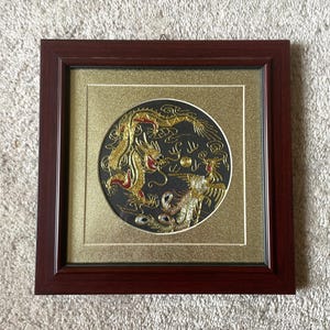 Può includere: Opera d'arte incorniciata con un disegno circolare di un drago e una fenice dorati su sfondo nero. L'opera è in una cornice di legno marrone scuro con un passe-partout dorato. Il drago e la fenice hanno dettagli dorati e rossi.