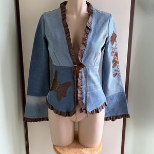 Puede incluir: Una chaqueta de mezclilla azul claro con ribetes de volantes marrones a lo largo de la abertura frontal y los puños. La chaqueta incluye una aplicación de mariposa marrón en el lado inferior derecho y bordados florales marrones en la manga izquierda.