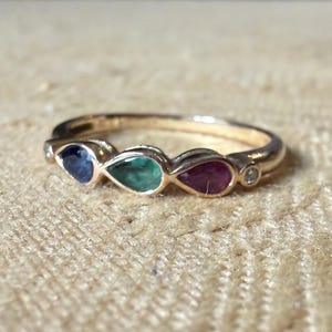 Puede incluir: Un anillo de oro con una hilera de tres piedras preciosas en forma de pera en azul, verde y rojo, flanqueadas por dos pequeñas piedras redondas transparentes. La banda es de oro pulido y las piedras están dispuestas horizontalmente.