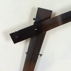 K&ouml;nnte beinhalten: Zwei schwarze Metallstreifen schneiden sich im rechten Winkel und bilden ein Kreuz auf wei&szlig;em Hintergrund. Die Streifen sind mit kleinen Schrauben am Hintergrund befestigt.