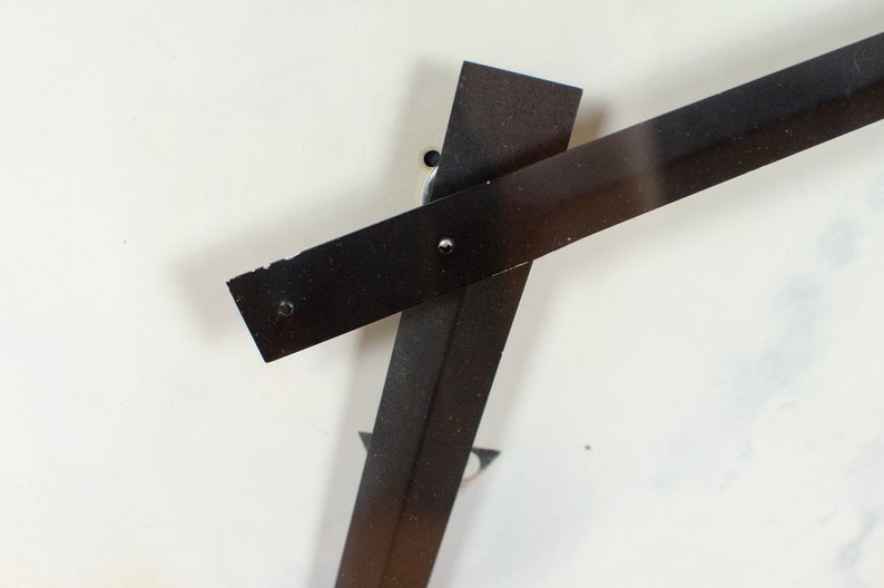 K&ouml;nnte beinhalten: Zwei schwarze Metallstreifen schneiden sich im rechten Winkel und bilden ein Kreuz auf wei&szlig;em Hintergrund. Die Streifen sind mit kleinen Schrauben am Hintergrund befestigt.