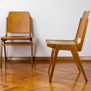 Può includere: Due sedie in legno con finitura marrone chiaro. Le sedie sono in compensato e hanno un design semplice e moderno. Le sedie sono su un pavimento in legno con un motivo a spina di pesce.