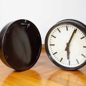 K&ouml;nnte beinhalten: Eine schwarze Vintage-Uhr mit wei&szlig;em Zifferblatt und schwarzen Zeigern. Die Uhr ist von ihrem R&uuml;cken getrennt, wodurch das Innenleben sichtbar wird.