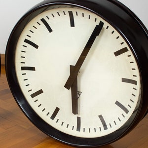 K&ouml;nnte beinhalten: Eine schwarze Uhr mit wei&szlig;em Zifferblatt und schwarzen Zeigern. Die Uhr hat eine schwarze L&uuml;nette und einen wei&szlig;en Hintergrund. Die Uhr befindet sich auf einer Holzoberfl&auml;che.