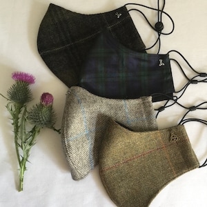 May include: Four face masks in different tweed patterns, including green, blue, and grey. Each mask has adjustable black ear loops and a decorative charm. A thistle flower is placed to the left of the masks.