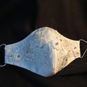 Könnte beinhalten: Weiße Gesichtsmaske mit weißer Spitze und floralen Verzierungen. Die Maske hat eine weiße Satinbasis und ist mit weißer Spitze und weißen Blumen verziert. Die Blumen haben kleine weiße Perlenakzente.